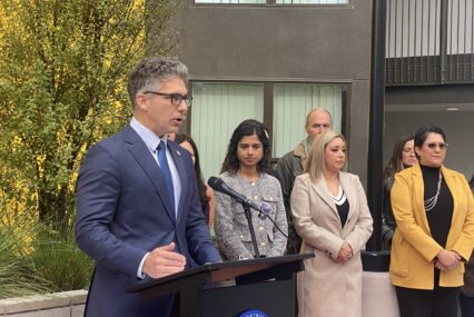 A man in a suit and tie speaks into a microphone at a podium, with people standing behind him