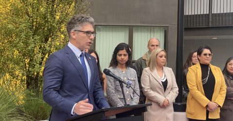 Un hombre con traje y corbata habla por un micrófono en un podio, con gente detrás de él.