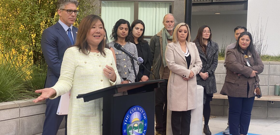 Một người phụ nữ đang phát biểu ngoài trời trên bục, phía sau là một nhóm người.