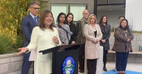 A woman speaks outside at a podium, with a group of people standing behind her