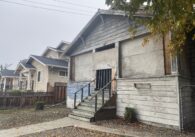 A boarded up wooden building next to a tan home.