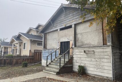 A boarded up wooden building next to a tan home.