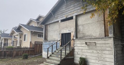 A boarded up wooden building next to a tan home.