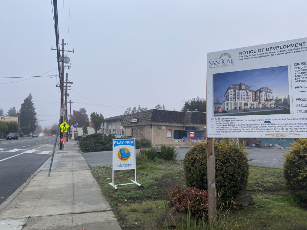 A sign on a grass field in front of a brown building next to a paved road.