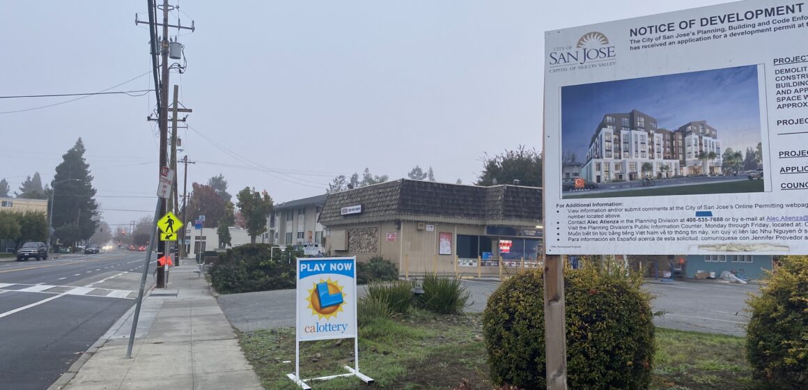 A sign on a grass field in front of a brown building next to a paved road.