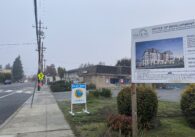 A sign on a grass field in front of a brown building next to a paved road.