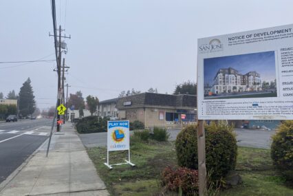 A sign on a grass field in front of a brown building next to a paved road.