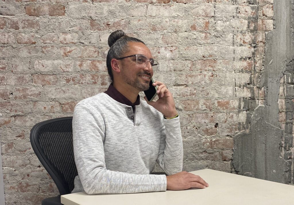 A man in a white sweater speaks on the phone at a desk in front of a brick wall.