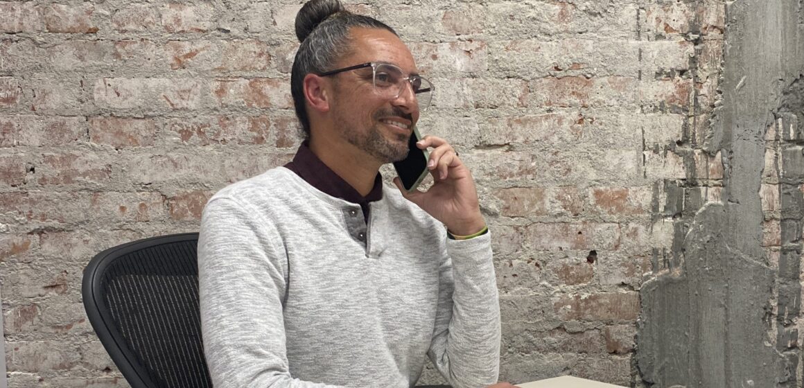 Gordon Chester A man in a white sweater speaks on the phone at a desk in front of a brick wall.