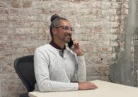 A man in a white sweater speaks on the phone at a desk in front of a brick wall.