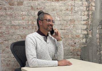 A man in a white sweater speaks on the phone at a desk in front of a brick wall.