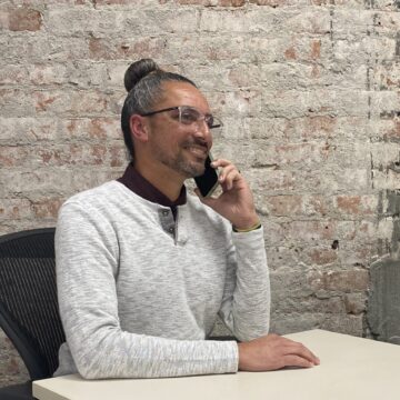 Gordon Chester A man in a white sweater speaks on the phone at a desk in front of a brick wall.