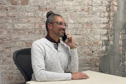 A man in a white sweater speaks on the phone at a desk in front of a brick wall.