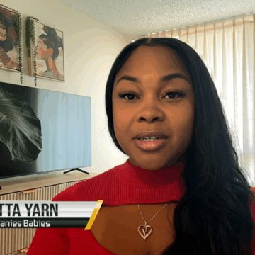 A Black woman sitting inside her home, with a plant, tv and blinds in the background