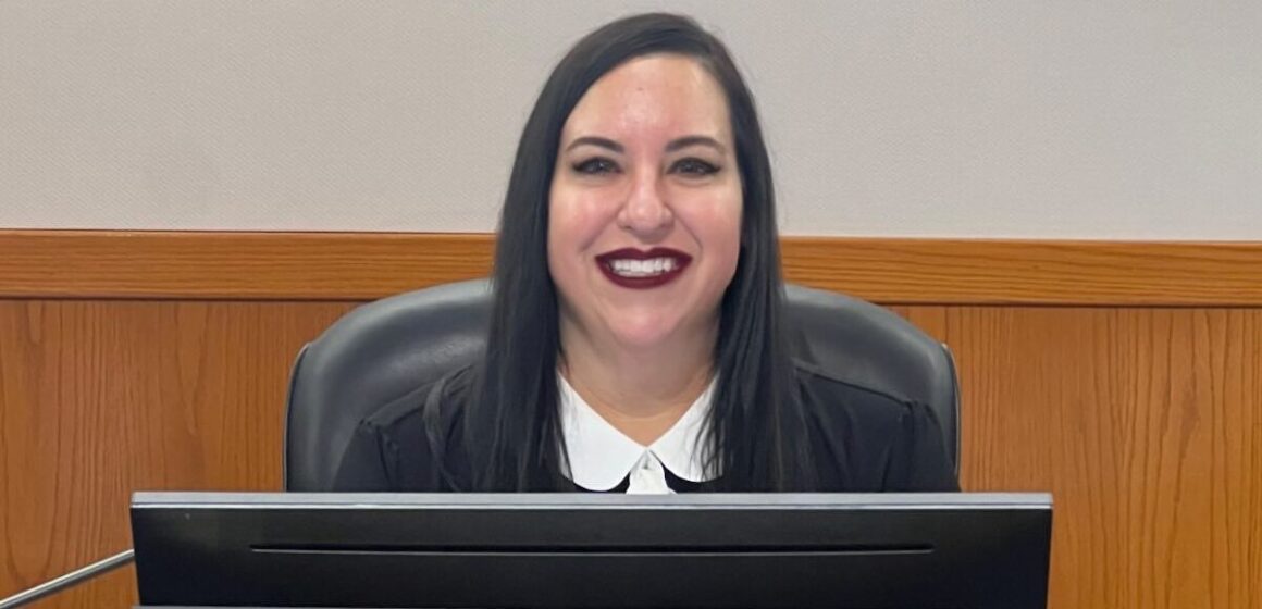 Aimee Escobar A woman in a black jacket sits behind a dais with a name plate.