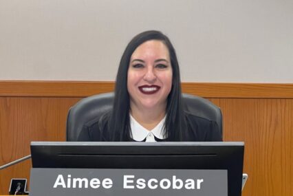 A woman in a black jacket sits behind a dais with a name plate.