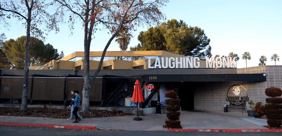 Ảnh chụp ngoại thất của một nhà hàng một tầng với biển hiệu lớn ghi "Laughing Monk" trên mái nhà, phía trước có vài cây xanh và biển hiệu neon "Open".