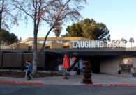 Photo of the exterior of a one-floor restaurant with a large sign reading "Laughing Monk" on top of the roof, with some trees in the front and a neon "Open" sign