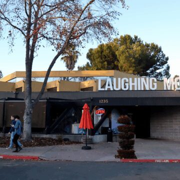Laughing Monk Brewery Sunnyvale Photo of the exterior of a one-floor restaurant with a large sign reading "Laughing Monk" on top of the roof, with some trees in the front and a neon "Open" sign