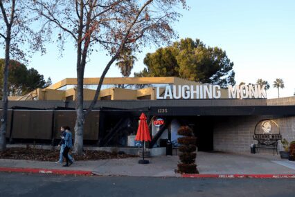 Photo of the exterior of a one-floor restaurant with a large sign reading "Laughing Monk" on top of the roof, with some trees in the front and a neon "Open" sign