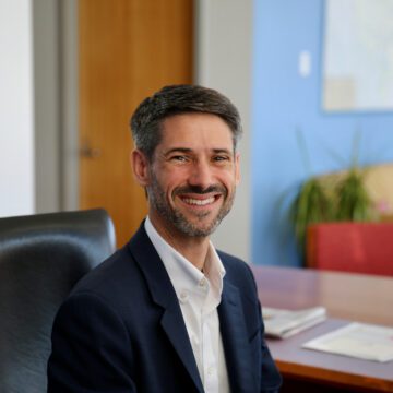 Mahan-Six-Months-2 A man in a suit jacket sitting at a desk