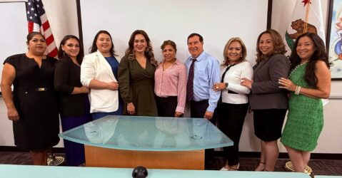 A row of people pose in front of a wall with flags in an office.