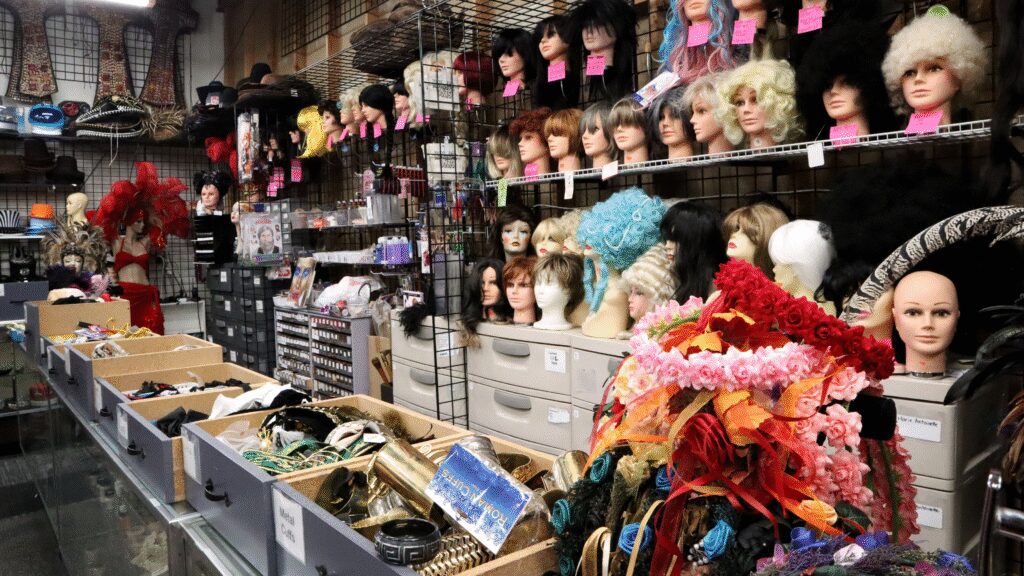 Photo of a store's counter, covered in pulled out drawers featuring different costume accessories, including a pile of flower crowns and metal arm cuffs. Behind the counter is a wall of wigs and shelves.