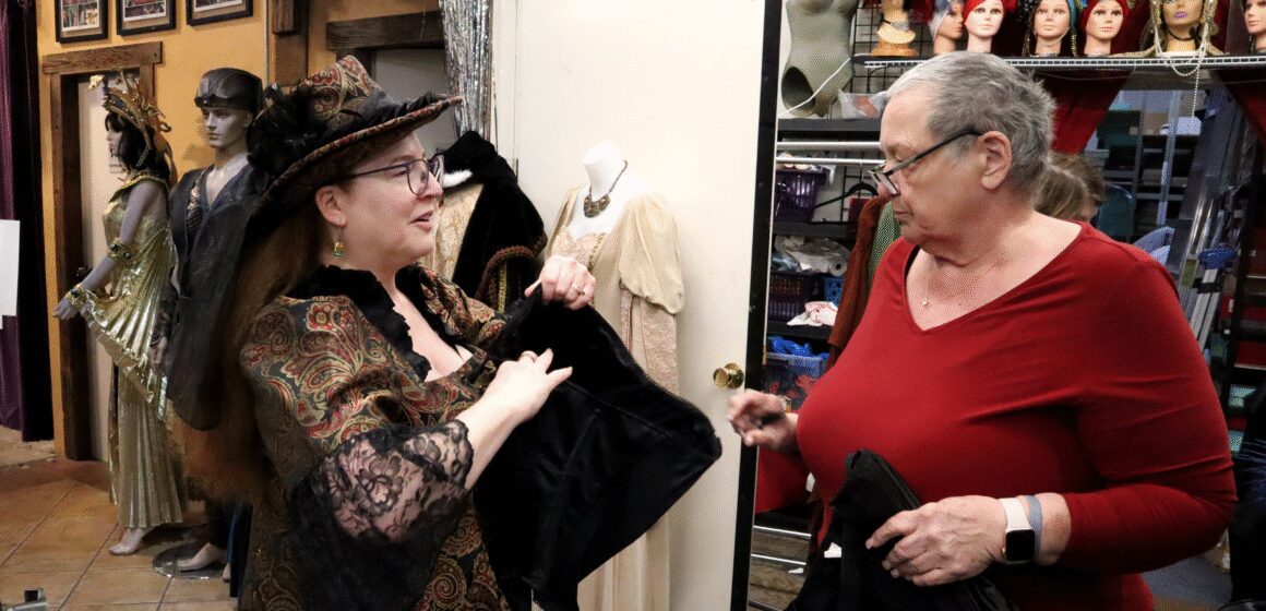 Photo of two women talking while standing in a cluttered costume shop. One woman is wearing a Victorian-inspired dress and head piece, holding a black corset, while the other woman inspects it.