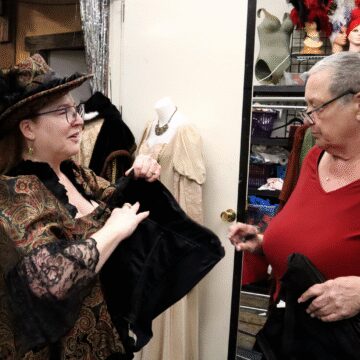 Photo of two women talking while standing in a cluttered costume shop. One woman is wearing a Victorian-inspired dress and head piece, holding a black corset, while the other woman inspects it.