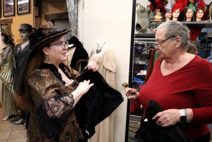 Photo of two women talking while standing in a cluttered costume shop. One woman is wearing a Victorian-inspired dress and head piece, holding a black corset, while the other woman inspects it.