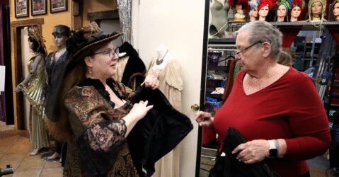 Photo of two women talking while standing in a cluttered costume shop. One woman is wearing a Victorian-inspired dress and head piece, holding a black corset, while the other woman inspects it.