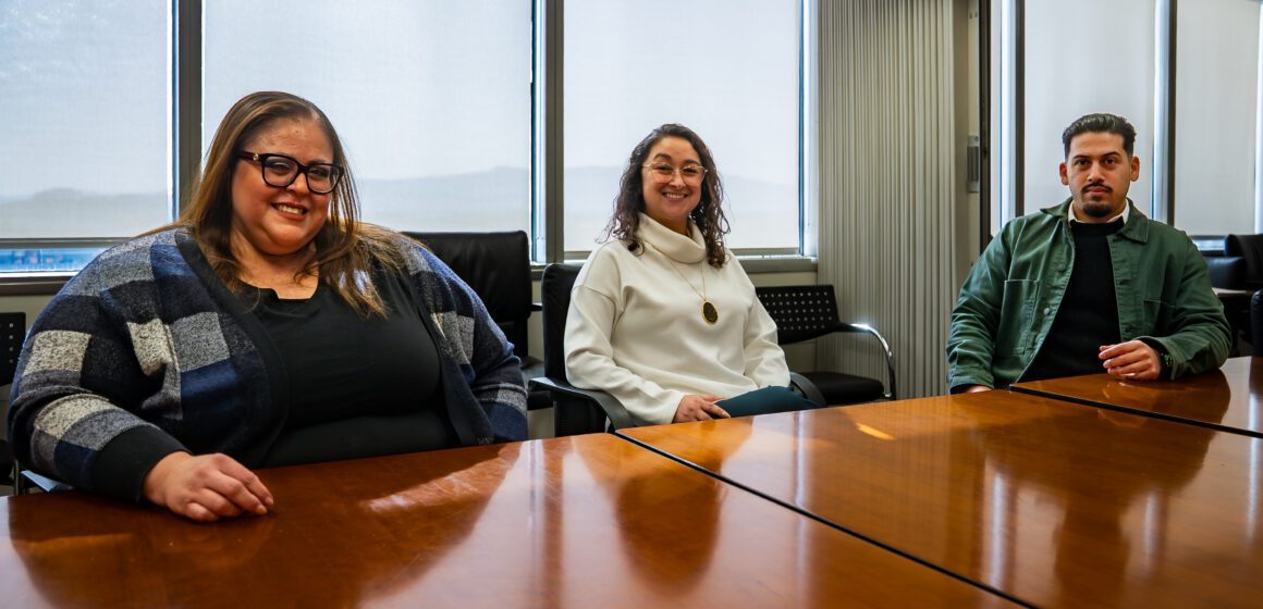 Three people sit at a conference table, with windows behind them