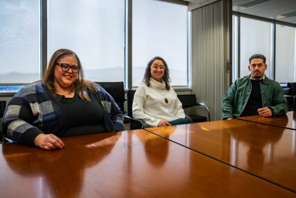 Three people sit at a conference table, with windows behind them