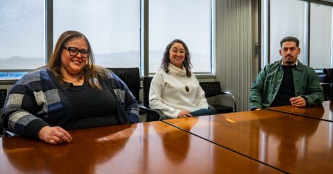 Three people sit at a conference table, with windows behind them