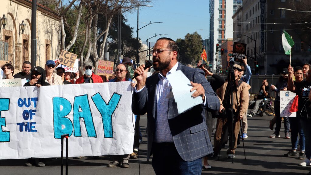 Photo of a man in a blue blazer holding a folder speaking on a microphone, with people around him holding signs reading "ICE OUT"