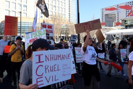 Photo of a group of people marching in front of the San Jose Convention Center, holding signs reading "INHUMANE CRUEL EVIL Due Process is American" and "LATINAS CONTRA ICE!!"