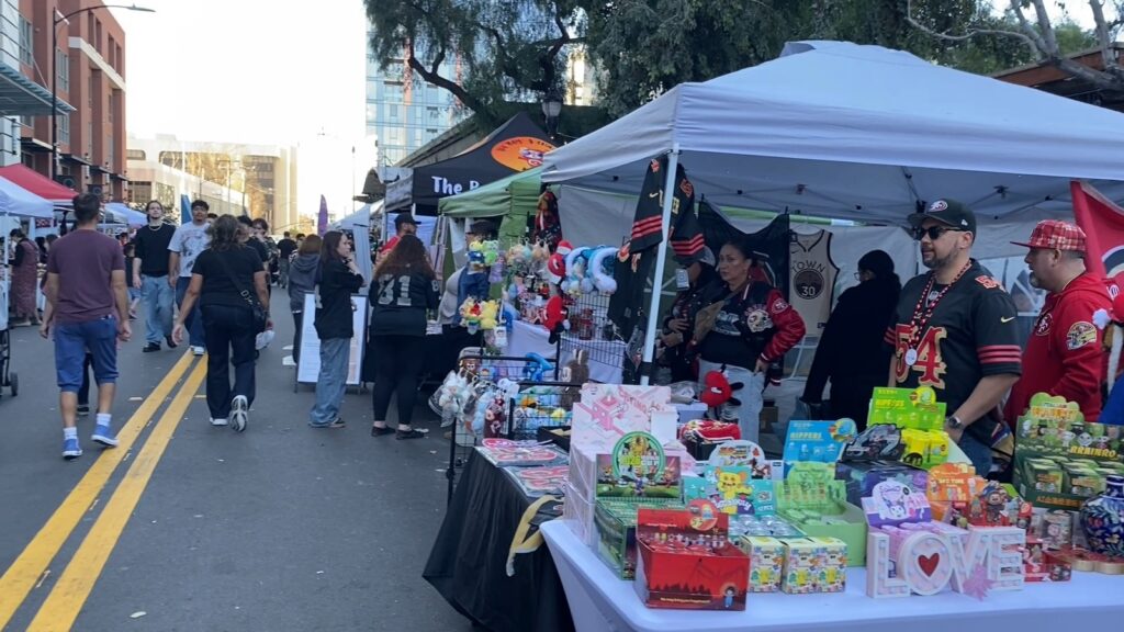 Vendors await customers underneath a pavilion. In front of them, passerby walk down a street lined by more vendor pavilions.