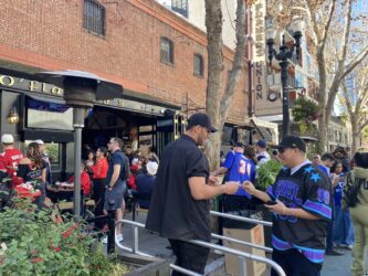 A man shows his ID to a bouncer to get into a bar. In the background, a large crowd is gathered.
