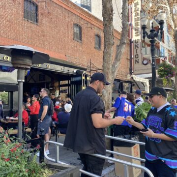 A man shows his ID to a bouncer to get into a bar. In the background, a large crowd is gathered.