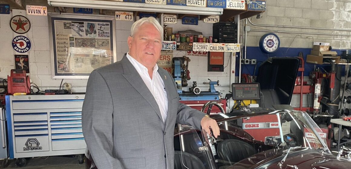 A man in a gray suit stands in front of a classic car.