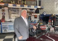 A man in a gray suit stands in front of a classic car.