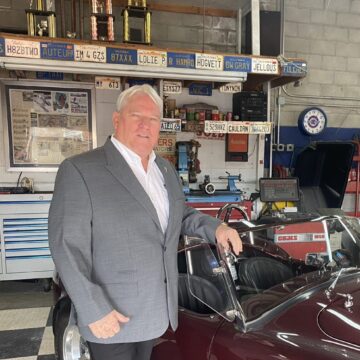 A man in a gray suit stands in front of a classic car.