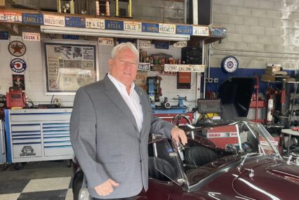 A man in a gray suit stands in front of a classic car.