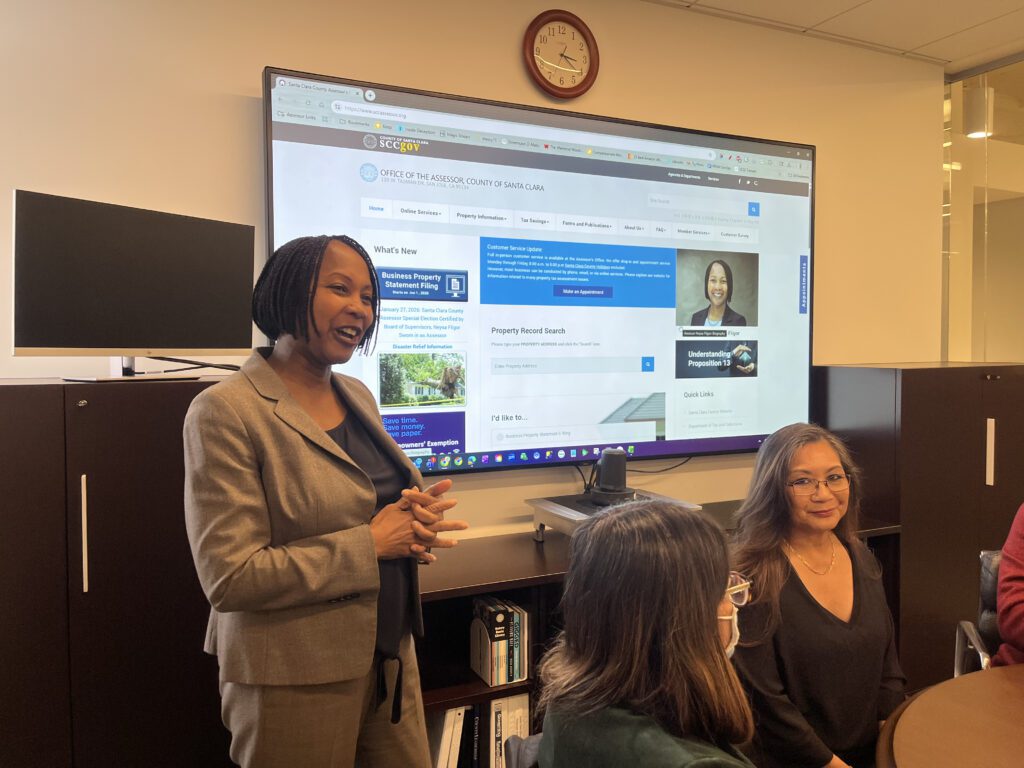 A Black woman in a suit standing in front of a projector screen talks with other people who are sitting down