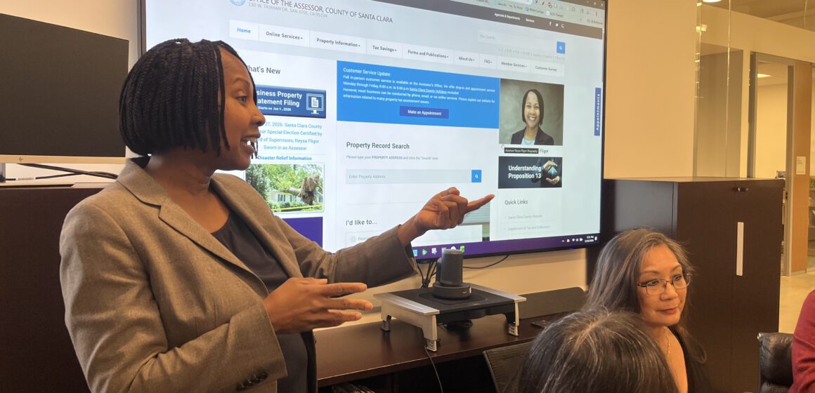 A Black woman in a suit standing in front of a projector screen talks with other people who are sitting down