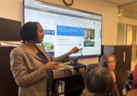 A Black woman in a suit standing in front of a projector screen talks with other people who are sitting down