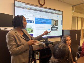 ASSESSOR A Black woman in a suit standing in front of a projector screen talks with other people who are sitting down