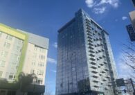 A glass high rise raises up into the sky across the street from a mid-rise apartment.