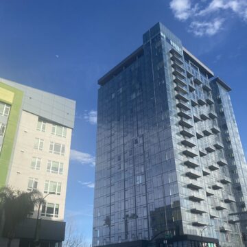 A glass high rise raises up into the sky across the street from a mid-rise apartment.