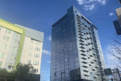 A glass high rise raises up into the sky across the street from a mid-rise apartment.